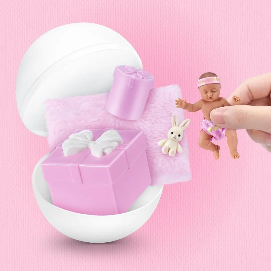 Cápsula de My Mini Baby Happy Birthday abierta por la mitad con la escena Gift Box, el conejito y el bebé sujetado entre dos dedos