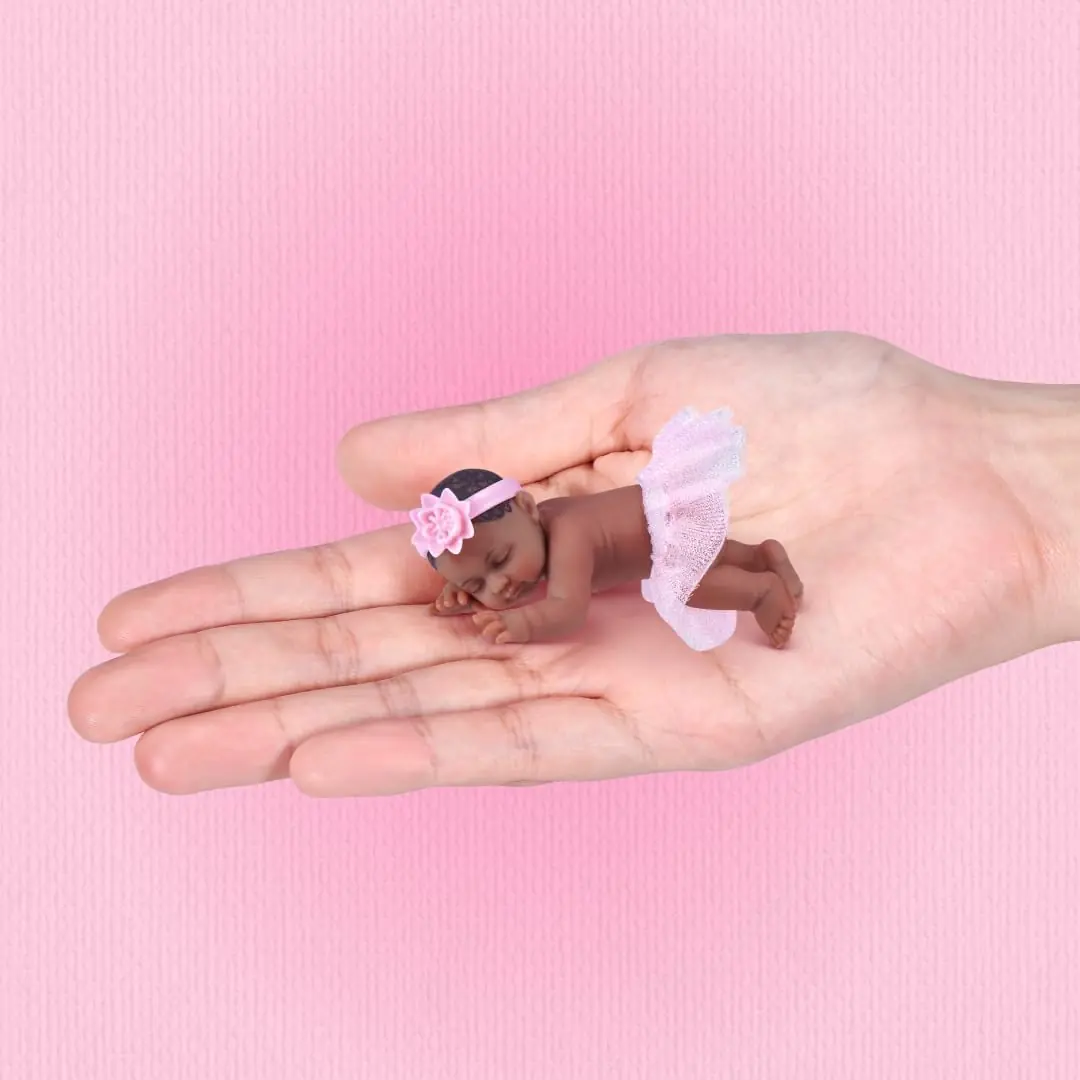 Un bebé de My Mini Baby Happy Birthday con tutú rosa y diadema de flor tumbado sobre la palma de una mano, mostrando su escala real