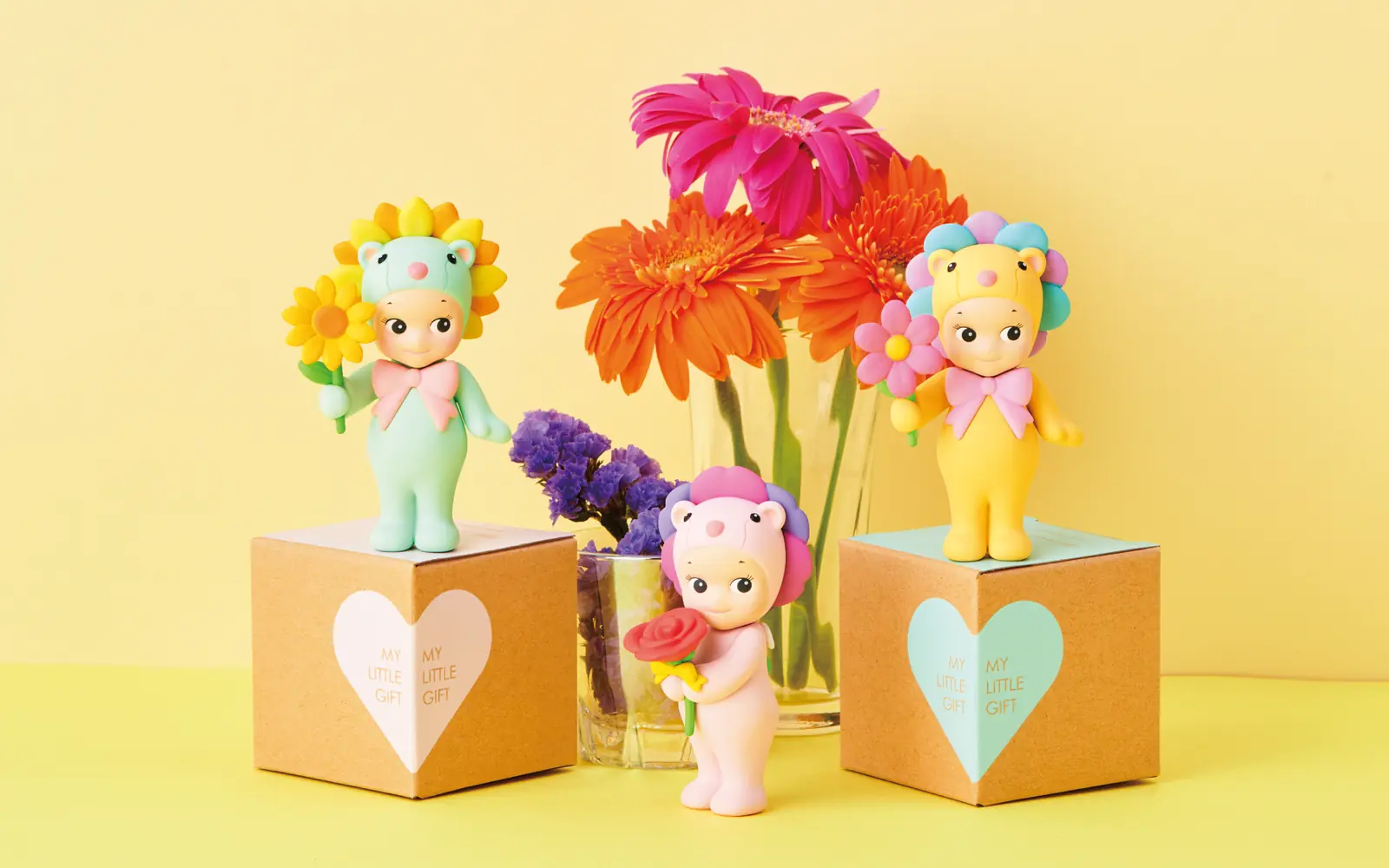 Tres figuras Sonny Angel Flower Gift con gerberas y cajas de regalo sobre fondo amarillo